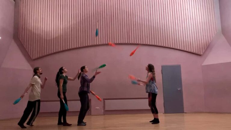 Four people juggling clubs in a large room with pink walls and a wooden floor. - Passing Zone