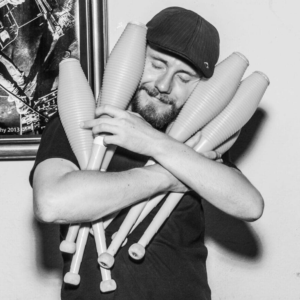 Man hugging juggling clubs, eyes closed, wearing a cap. Juggler with props. - Passing Zone