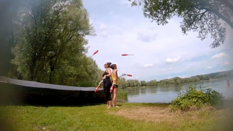 Two women juggling clubs by a lake; outdoor recreational activity. - Passing Zone