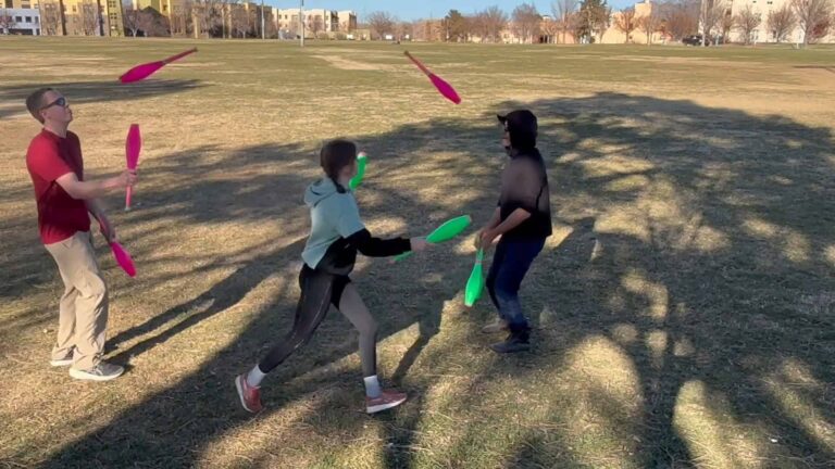 Three people juggling colorful clubs in a grassy park with buildings in the background. - Passing Zone