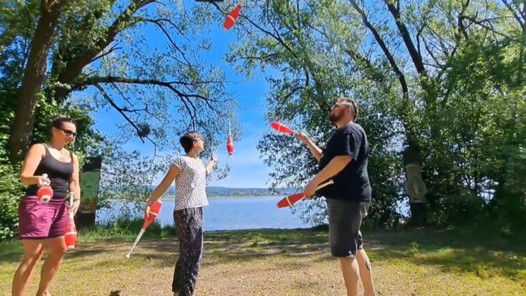 Three people juggling red clubs outdoors near a lake. Mini Chopabout No. 3. - Passing Zone