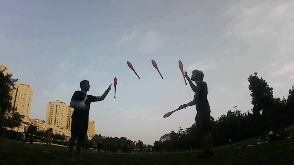 Two jugglers perform a 3-handed siteswap with clubs in a park setting with buildings in the background. - Passing Zone