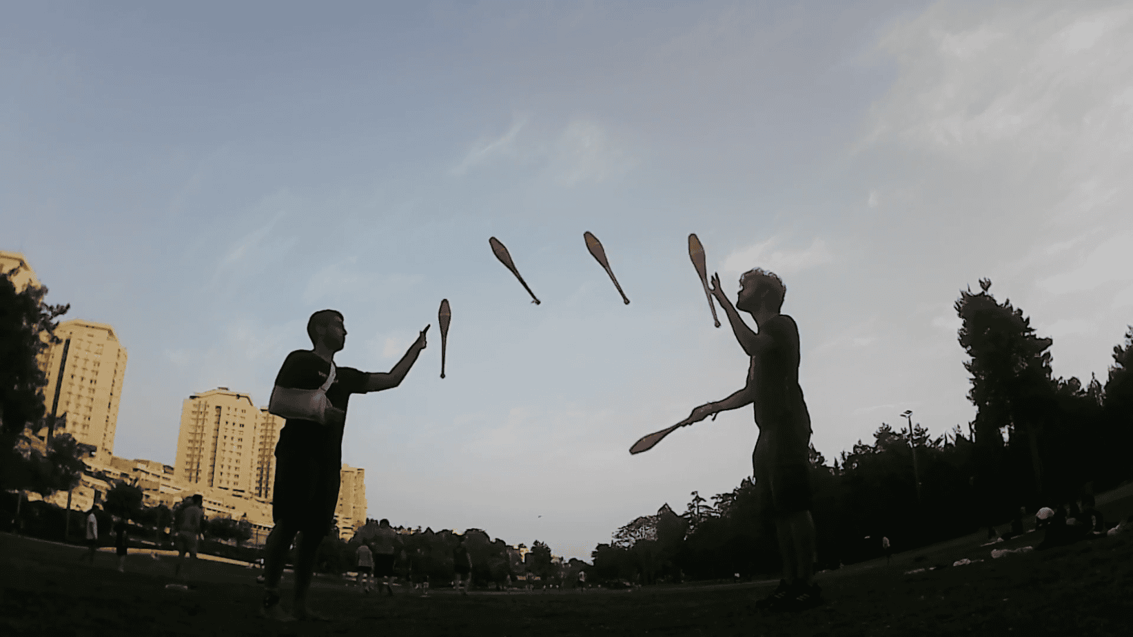 Two jugglers perform a 3-handed siteswap with clubs in a park setting with buildings in the background. - Passing Zone
